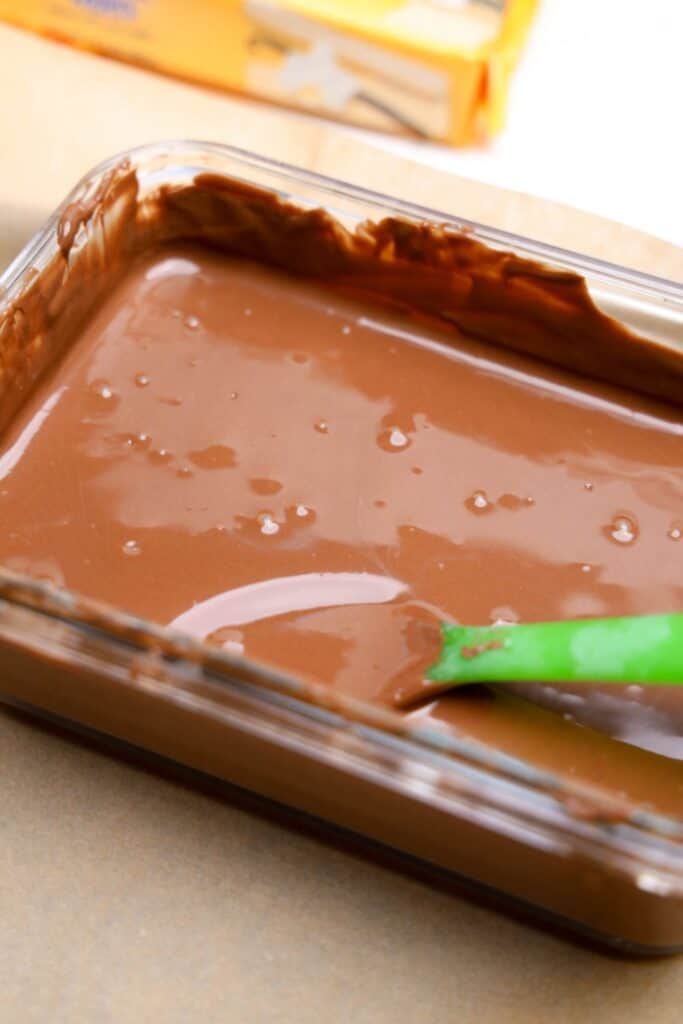 Glass dish filled with smooth, melted milk chocolate, being stirred with a green spatula. The chocolate is glossy and ready for dipping cookies, with a box of vanilla wafers blurred in the background.