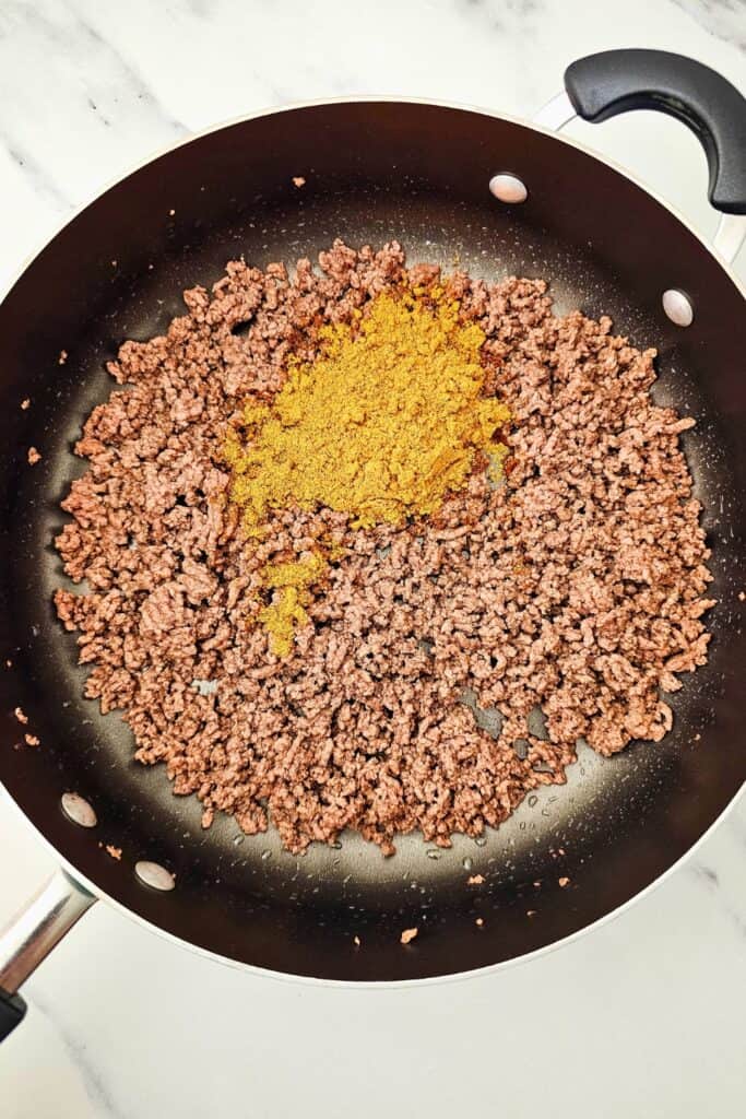 Cooked ground beef in a nonstick skillet with a generous pile of taco seasoning sprinkled on top, ready to be mixed in. This seasoned beef mixture forms the flavorful base for Tex-Mex sheet pan quesadillas.