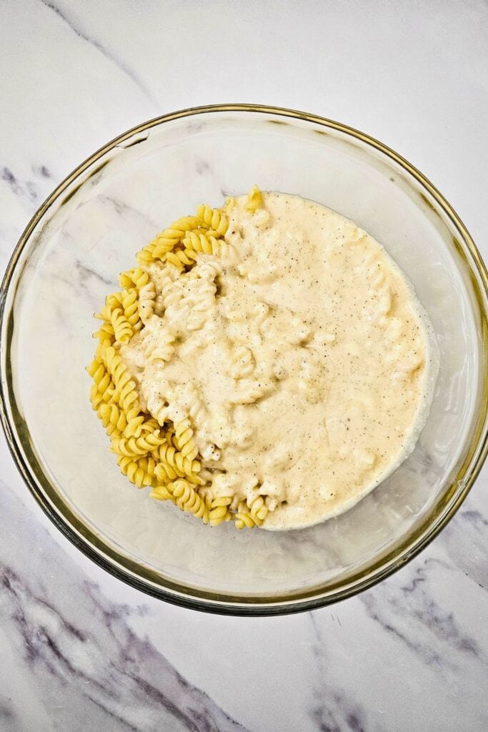 Glass mixing bowl containing cooked rotini pasta partially covered with a creamy mayonnaise-based dressing, ready to be mixed together for pasta salad.