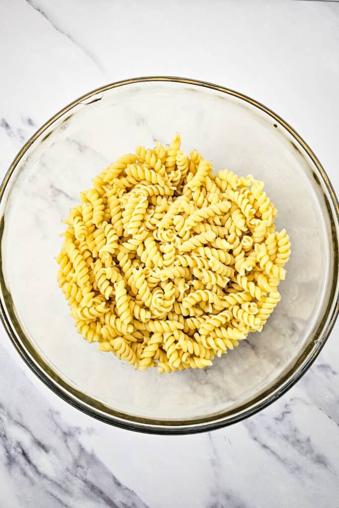 Overhead view of cooked rotini pasta in a large clear glass mixing bowl, placed on a white marble surface. The pasta is drained and ready for salad preparation.