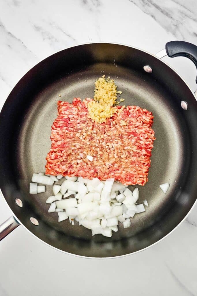 Raw Italian sausage with diced onion and minced garlic in a large skillet. The ingredients are ready to cook together for the sheet pan lasagna base.
