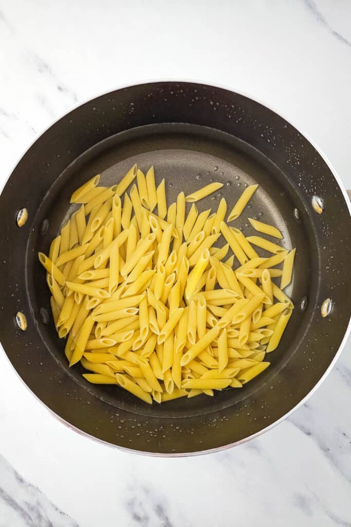 Uncooked penne pasta sitting in a black pot with a thin layer of water at the bottom, ready to be boiled. The pot is placed on a white marble countertop, capturing an early step in the pasta salad preparation process.