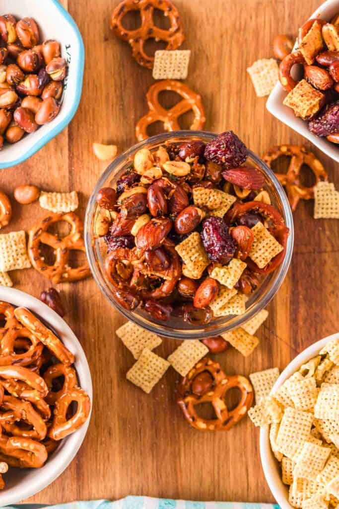 A bowl of cool ranch jerky trail mix filled with seasoned nuts dried cranberries pretzels and square cereal pieces placed on a wooden surface. Additional bowls and scattered ingredients surround it highlighting the mix of textures.