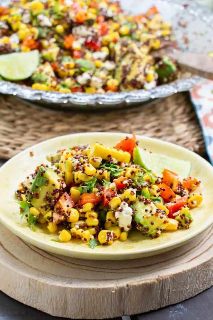 Corn avocado salad made with sweet corn, avocado chunks, diced red bell pepper, quinoa, fresh herbs, and crumbled cheese tossed together in a light dressing. A lime wedge sits beside the colorful salad, highlighting the fresh ingredients and vibrant texture of the dish.