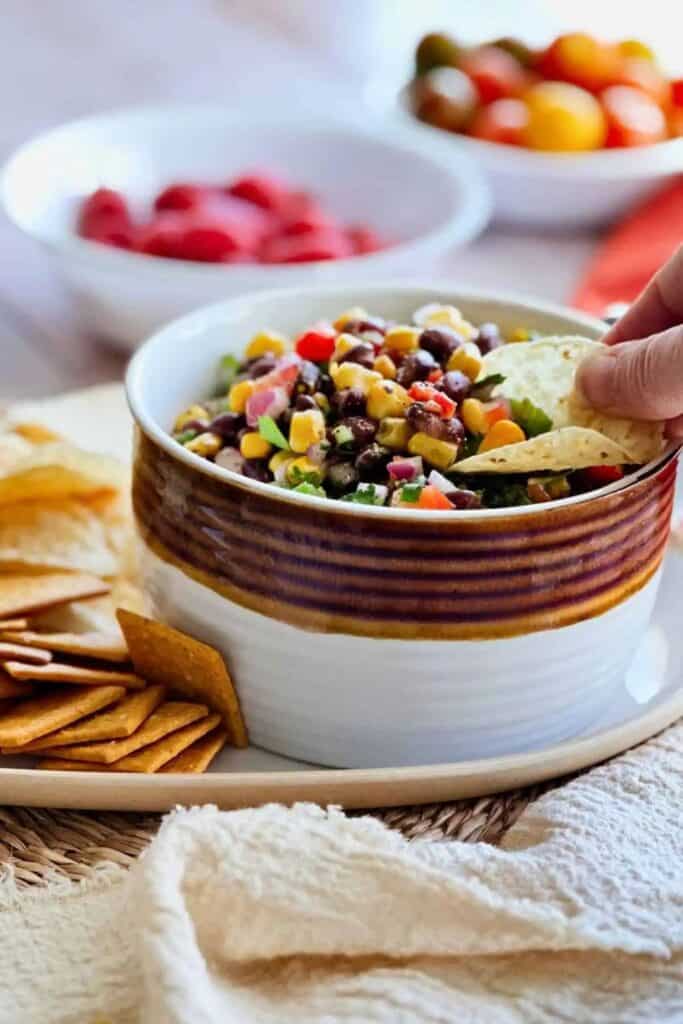 A ceramic bowl filled with colorful corn and black bean salsa, featuring diced red onion, tomatoes, and cilantro, with a tortilla chip being dipped in—served on a tray alongside crackers and chips, with bowls of fresh ingredients blurred in the background.