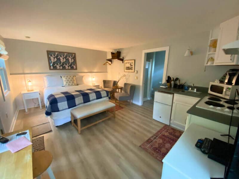 Cozy studio-style cottage with a king bed dressed in a navy and white checkered blanket, a small dining area, and a kitchenette with a stovetop, sink, and microwave. The light wood floors and soft lighting create a warm, pet-friendly retreat in Bar Harbor, Maine.
