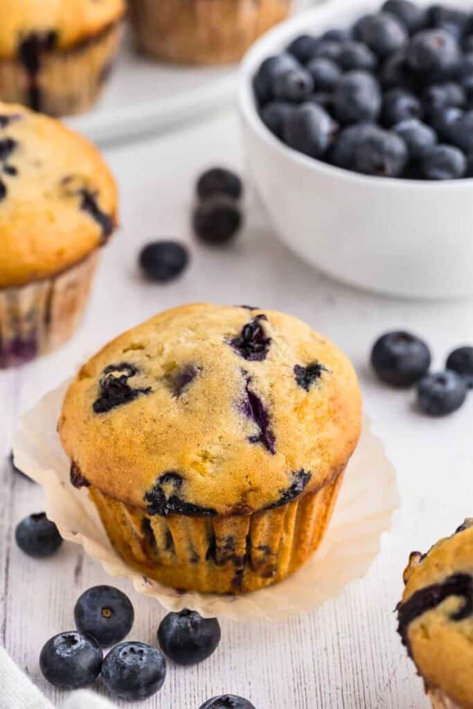 Close up of blueberry cottage cheese muffins with a golden brown domed top and juicy blueberries baked throughout. The tender crumb appears moist and fluffy, with fresh blueberries scattered around and a bowl of berries in the background. 