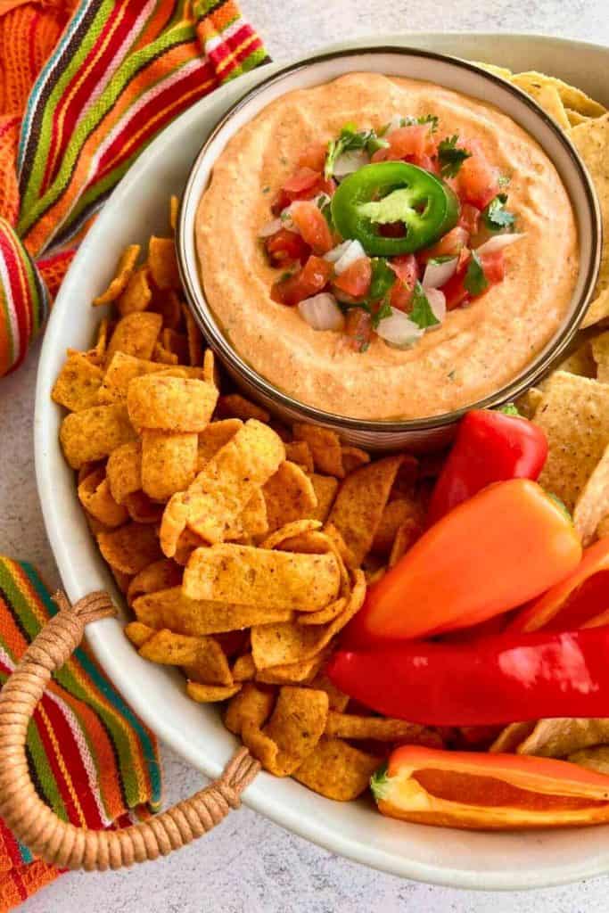 Bowl of creamy Cottage Cheese Taco Dip topped with fresh pico de gallo and a sliced jalapeño, surrounded by Fritos, tortilla chips, and sliced mini bell peppers for dipping. The dish is served in a large platter with a colorful striped napkin adding a festive, Southwestern touch.