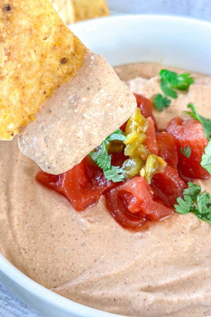 Creamy cottage cheese taco dip in a white bowl, topped with diced tomatoes, green chiles, and fresh cilantro, with a tortilla chip dipped in the center. A smooth and tangy dip for football tailgate parties.