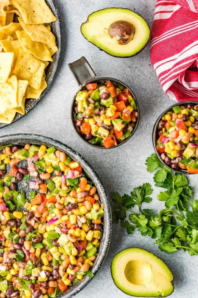 Colorful cowboy caviar served in a large bowl and smaller cups, made with black beans, corn, bell peppers, avocado, tomatoes, red onion, and cilantro. Surrounding the dip are tortilla chips, fresh avocado halves, and sprigs of cilantro on a light gray surface.