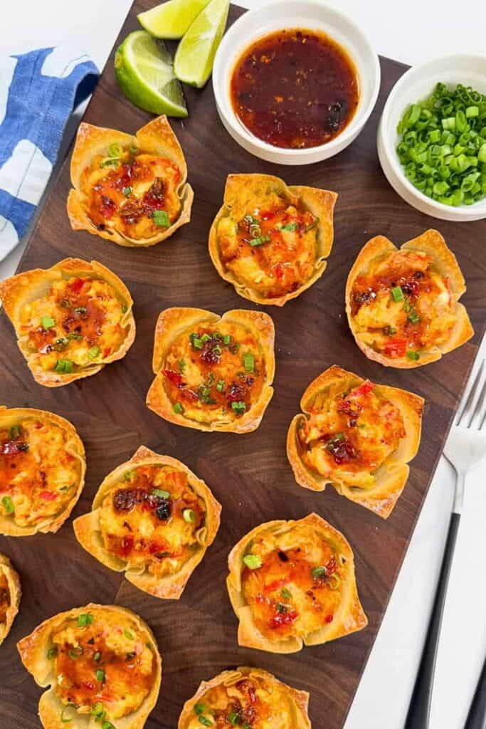 A wooden serving board covered with crispy golden Crab Rangoon Wonton Cups filled with creamy crab and red pepper filling, drizzled with sweet chili sauce and sprinkled with sliced green onions, with bowls of extra sauce and green onions and fresh lime wedges alongside.