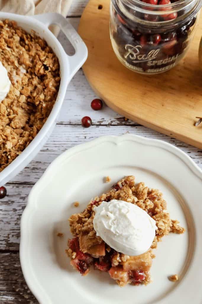 A serving of warm apple cranberry crisp topped with a scoop of vanilla ice cream on a white plate. The dessert features a golden oat crumble and is shown next to a baking dish and a mason jar filled with fresh cranberries on a rustic wooden surface.