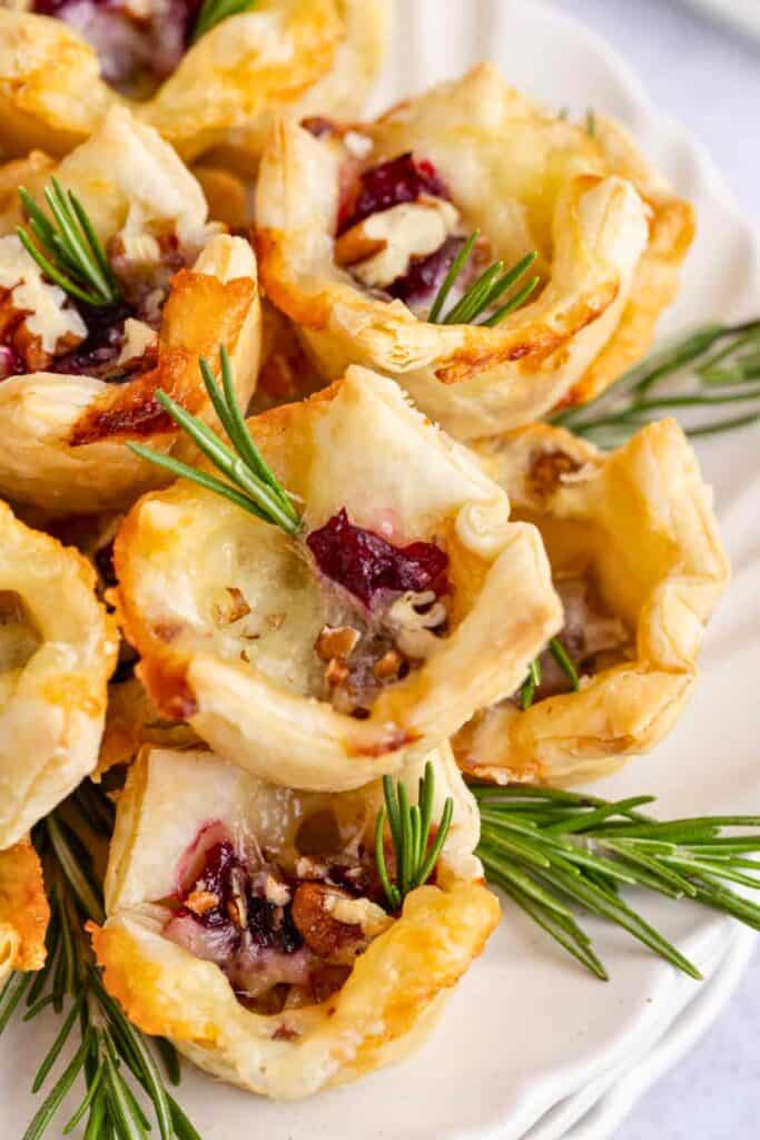 Cranberry Brie bites made with golden puff pastry cups filled with melted Brie cranberry sauce and chopped pecans. Fresh rosemary sprigs garnish the bite sized appetizers on a white serving platter.