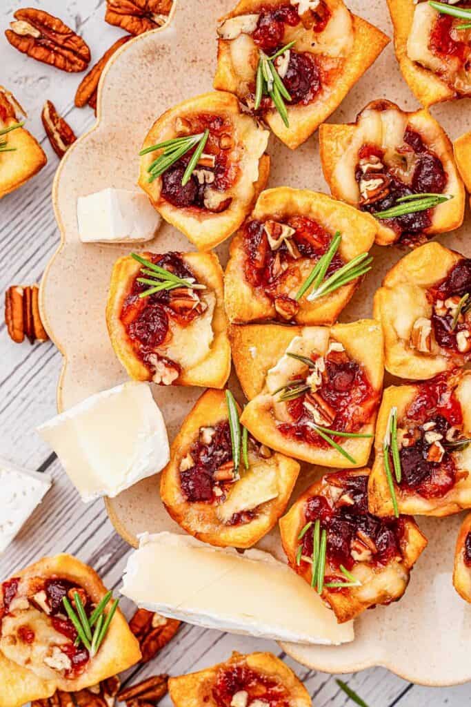 An overhead view of cranberry brie bites arranged on a scalloped serving platter with wedges of brie and scattered pecans around them. The golden pastry cups are filled with melted brie and cranberry sauce and topped with chopped pecans and fresh rosemary.