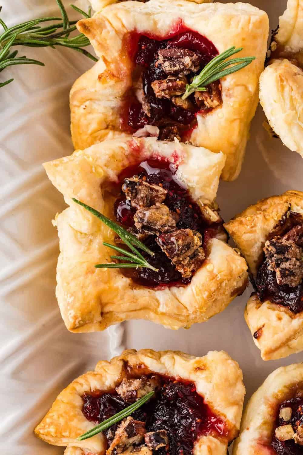 Overhead view of 3 Golden puff pastry Cranberry Brie Bites filled with creamy brie and cranberry sauce, topped with chopped pecans and fresh rosemary on a patterned plate.