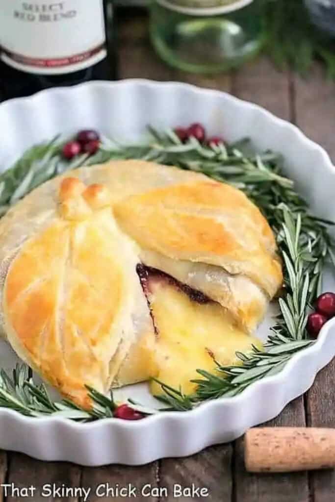 Cranberry Brie en Croute with melted Brie and cranberry filling wrapped in golden brown puff pastry with a wedge cut out. The baked Brie is served in a white dish surrounded by fresh rosemary sprigs and cranberries.