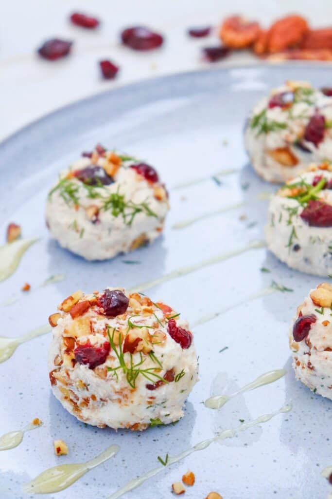 A close up of cranberry goat cheese bites arranged across a pale blue serving platter. The creamy white bites are packed with chopped pecans and dark red dried cranberries and topped with fresh green dill while thin streaks of golden honey are drizzled across the plate.