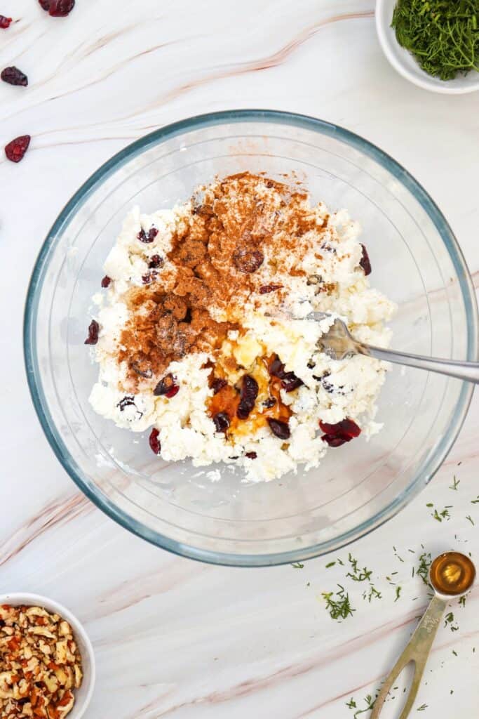 Process shot showing a cranberry cheese mixture in a clear glass bowl topped with cinnamon and a drizzle of honey before mixing. Dried cranberries are scattered throughout the cheese mixture, with chopped pecans, fresh dill, and a measuring spoon arranged nearby.