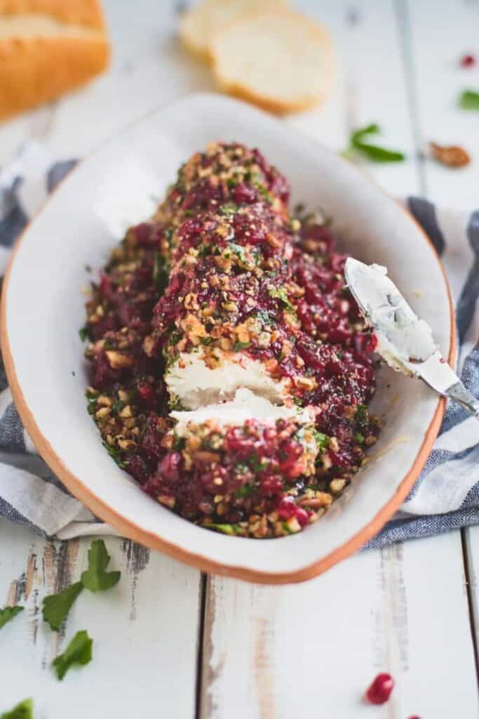 A cranberry goat cheese log coated with cranberry sauce, chopped nuts, and fresh herbs sits in a white serving dish. A portion has been sliced away to reveal the creamy goat cheese center, with a cheese knife and slices of bread nearby for serving.