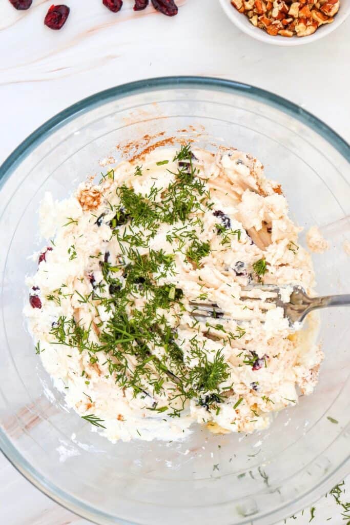 Process shot showing chopped fresh dill sprinkled over a creamy goat cheese mixture with dried cranberries and cinnamon, vanilla and honey in a clear glass bowl. A fork rests in the partially mixed filling, while a bowl of chopped pecans and several dried cranberries sit nearby.