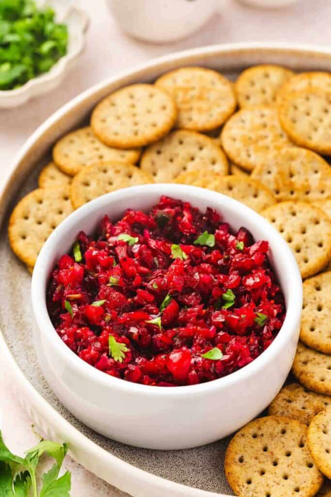 A bowl of bright red cranberry jalapeño salsa topped with fresh cilantro sits in the center of a serving platter. Round crackers are arranged around the bowl for scooping up the chunky cranberry salsa.