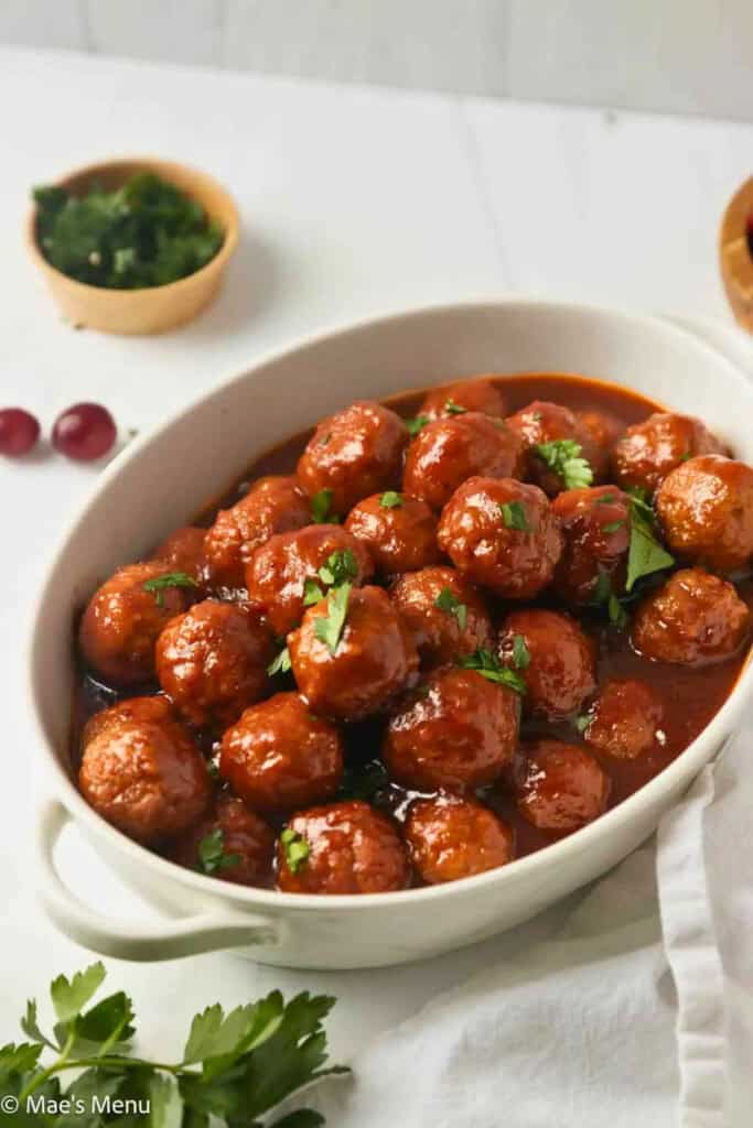 A white serving dish filled with glossy cranberry meatballs in a rich red sauce, topped with fresh chopped parsley are an easy Christmas potluck idea.