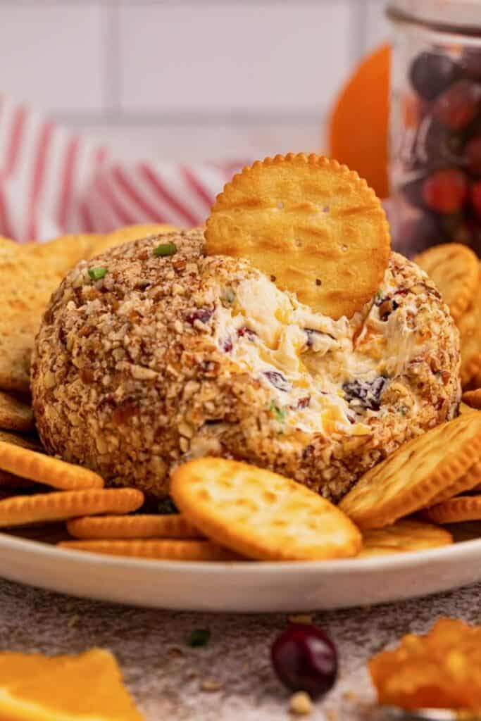 A cranberry orange cheese ball coated with chopped pecans sits in the center of a platter surrounded by round crackers. A cracker is dipped into the creamy cheese ball, revealing bits of dried cranberries, orange cheese, and green onions inside.