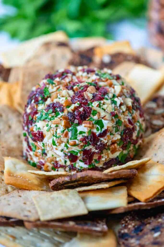 A cranberry pecan cheese ball coated with chopped herbs and nuts and surrounded by crisp crackers for an easy Thanksgiving appetizer.