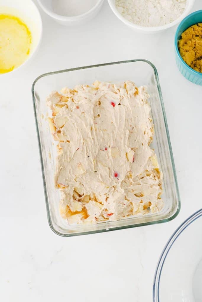 A glass baking dish filled with a creamy apple dip mixture, featuring visible chunks of fresh apples and swirls of spiced cream cheese. Surrounding the dish are bowls of melted butter, white sugar, flour, and brown sugar, set on a white countertop.