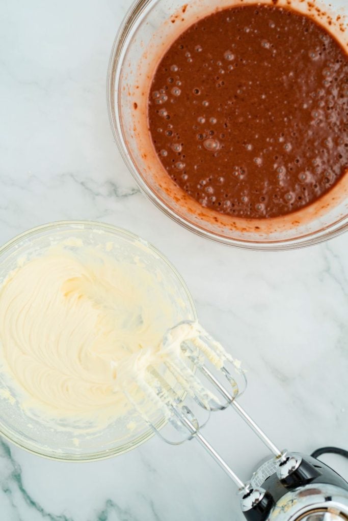 Two glass mixing bowls on a marble countertop—one with a creamy, pale yellow mixture of butter, cream cheese, and sugar, and the other with a smooth chocolate pudding mixture, both prepared with an electric hand mixer.