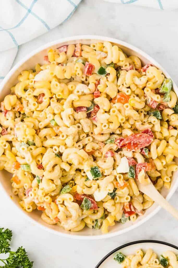 A large white bowl filled with creamy Amish macaroni salad, made with elbow macaroni, diced red and green bell peppers, chopped hard-boiled eggs, and a rich mayo-based dressing. A wooden spoon rests in the bowl, ready for serving.