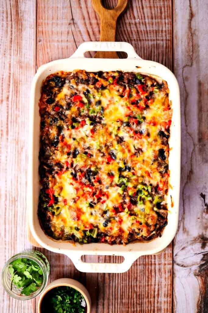 Creamy burrito casserole topped with melted cheese, black beans, diced red and green peppers, and fresh herbs in a rectangular baking dish.