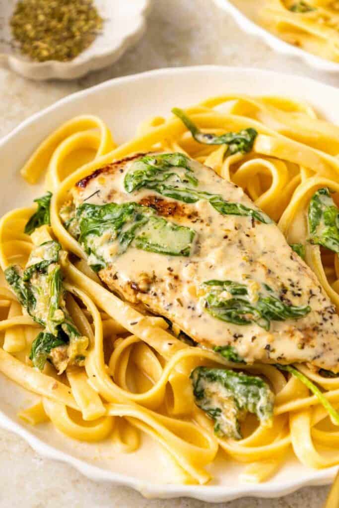 Creamy chicken and spinach served over wide fettuccine noodles with a seared chicken breast coated in a rich garlic cream sauce. Close up of this one pan dinner plated with tender pasta and wilted spinach with a small dish of herbs blurred in the background.