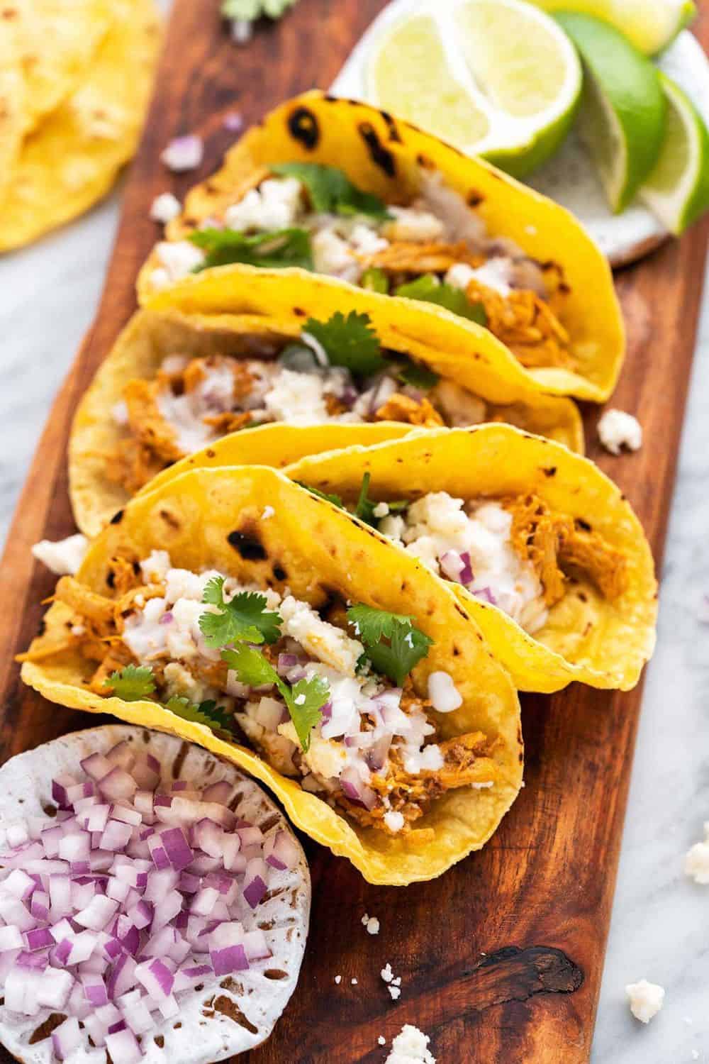 Four crispy corn tortillas filled with shredded chipotle chicken, crumbled queso fresco, diced red onion, fresh cilantro, and a drizzle of creamy sauce, served with lime wedges on a wooden board. These chipotle pulled chicken tacos are a flavorful and easy meal with smoky, tangy toppings.