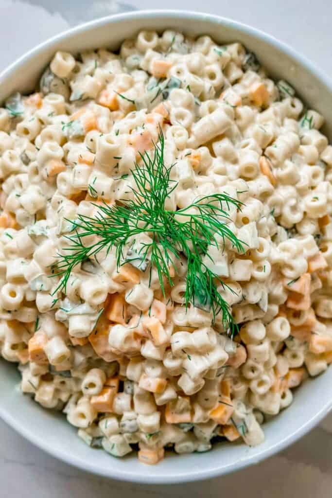 Creamy dill pickle cold pasta salad served in a white bowl, made with ditalini pasta, diced cheddar cheese, chopped pickles, and fresh dill. The salad is coated in a rich, tangy dressing and topped with a sprig of dill for garnish.
