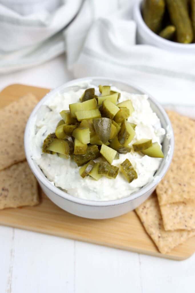 A white bowl filled with creamy dill pickle dip, generously topped with chopped pickles, sits on a wooden board. Whole grain crackers surround the dip, ready for scooping.