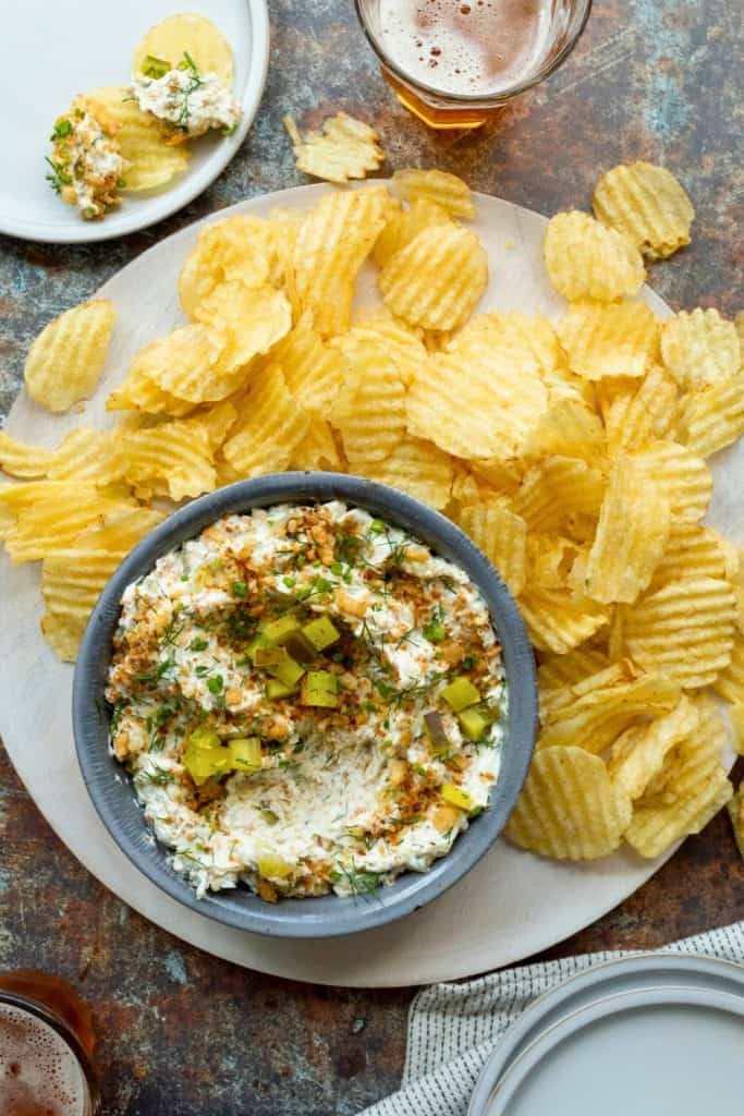 Creamy fried dill pickle dip served in a gray bowl, topped with chopped pickles, herbs, and crispy golden crumbs. Surrounded by ridged potato chips on a round wooden board, with a glass of beer nearby for a perfect snack pairing.