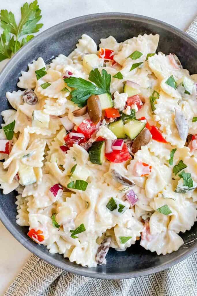 Creamy Greek tzatziki pasta salad served in a dark bowl, made with bowtie pasta, diced cucumbers, tomatoes, red onions, kalamata olives, and crumbled feta cheese, all coated in a rich tzatziki-style dressing. Garnished with fresh parsley for a refreshing Mediterranean touch.