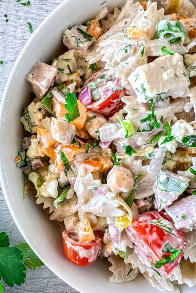 Close-up of a cold bowtie pasta salad in a white bowl, featuring creamy farfalle pasta mixed with chickpeas, chicken, ham, cherry tomatoes, diced bell peppers, red onion, celery, and herbs. The vibrant ingredients are coated in a rich, herbed dressing.