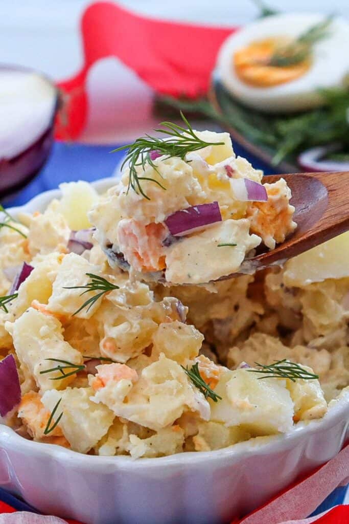 Creamy potato salad served in a white dish, topped with fresh dill, red onion, and chunks of carrot. A wooden spoon lifts a hearty scoop, with a sliced boiled egg and herbs blurred in the background