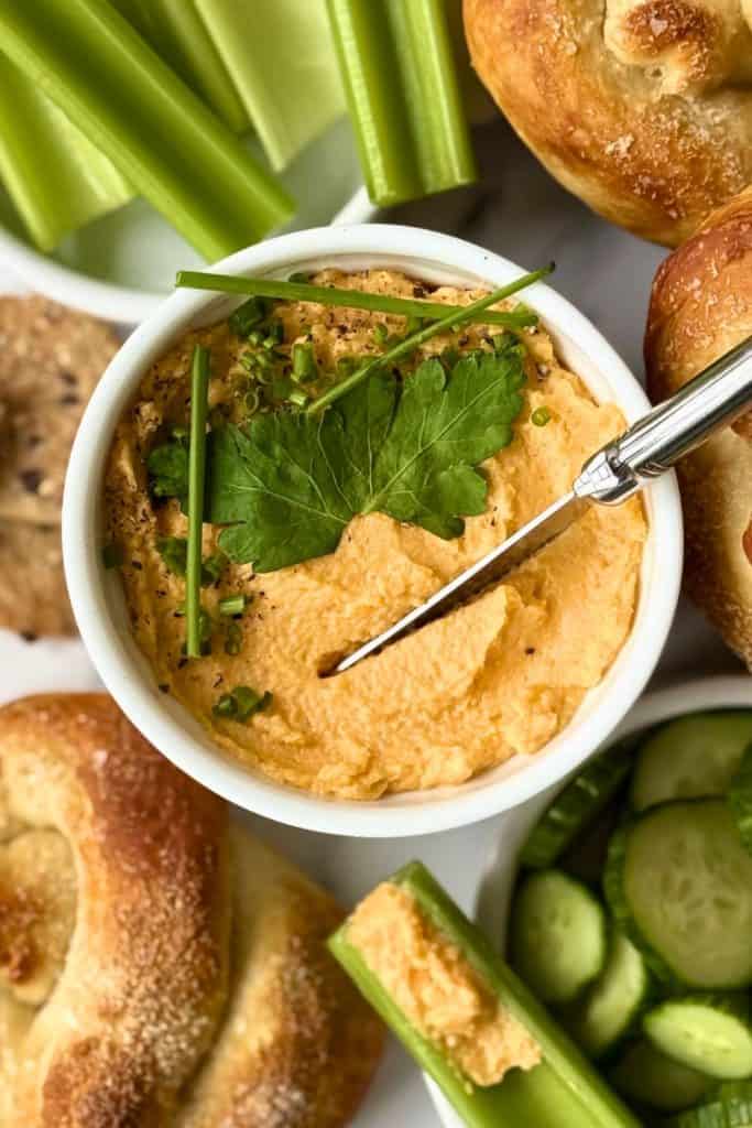Creamy Pub Style Beer Cheese Dip in a white ramekin, garnished with fresh parsley, chopped chives, and cracked black pepper. Surrounded by soft pretzels, celery sticks, sliced cucumbers, and crackers.