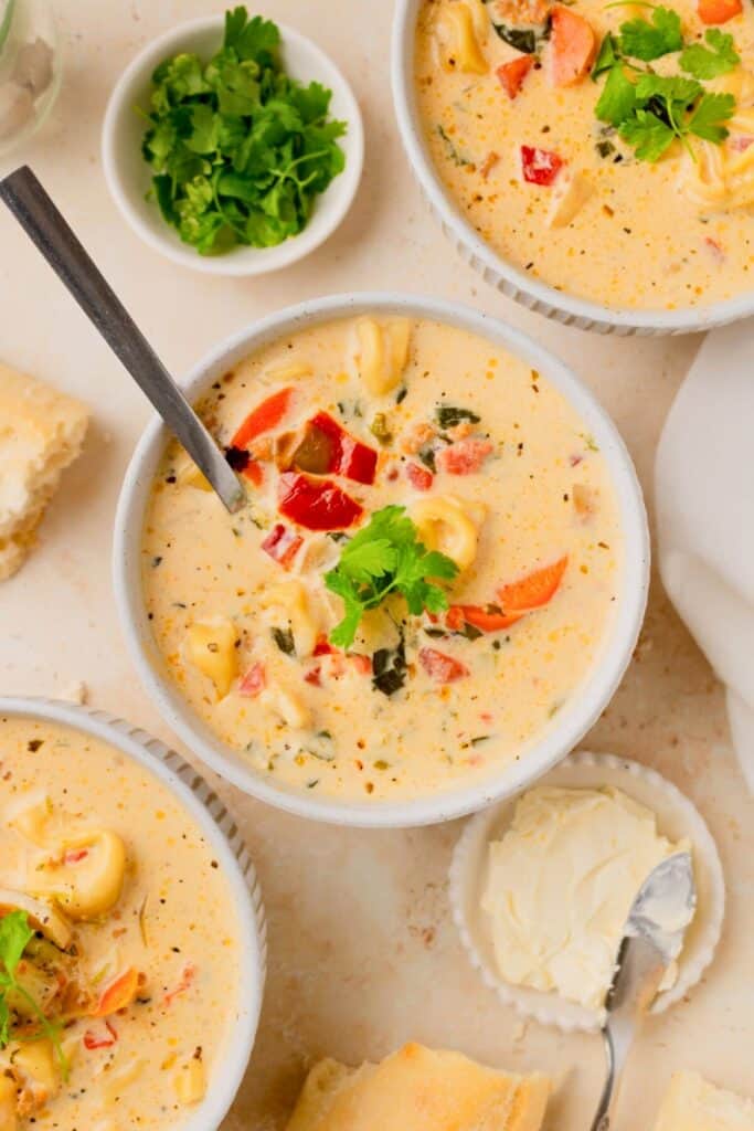 Creamy sausage and tortellini soup with red peppers and fresh herbs is served in a white bowl. Crusty bread and butter sit alongside the rich creamy soup.