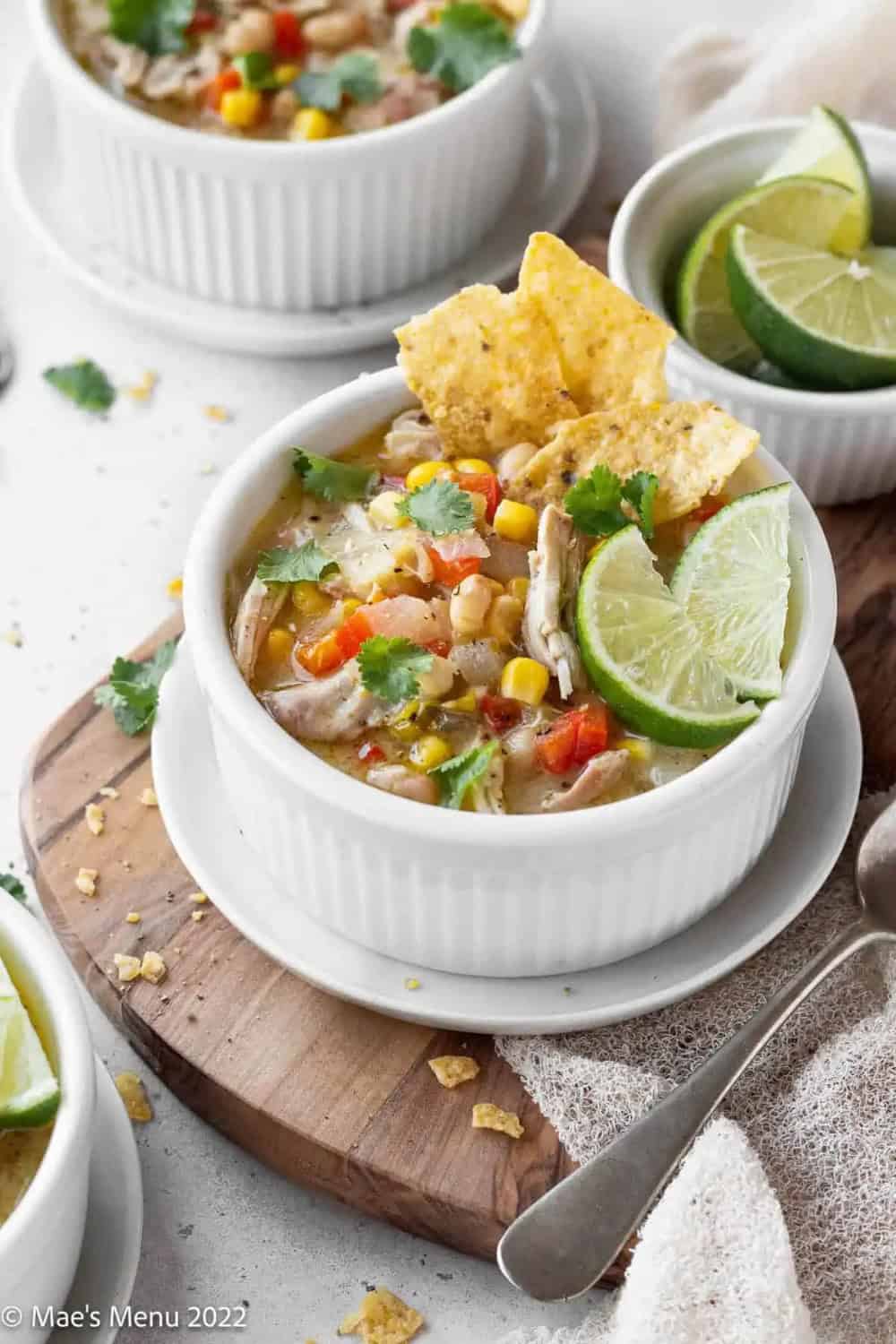 Creamy white tortilla soup with shredded chicken corn and red peppers is served in a white bowl. The soup is topped with crispy tortilla chips fresh cilantro and lime wedges.