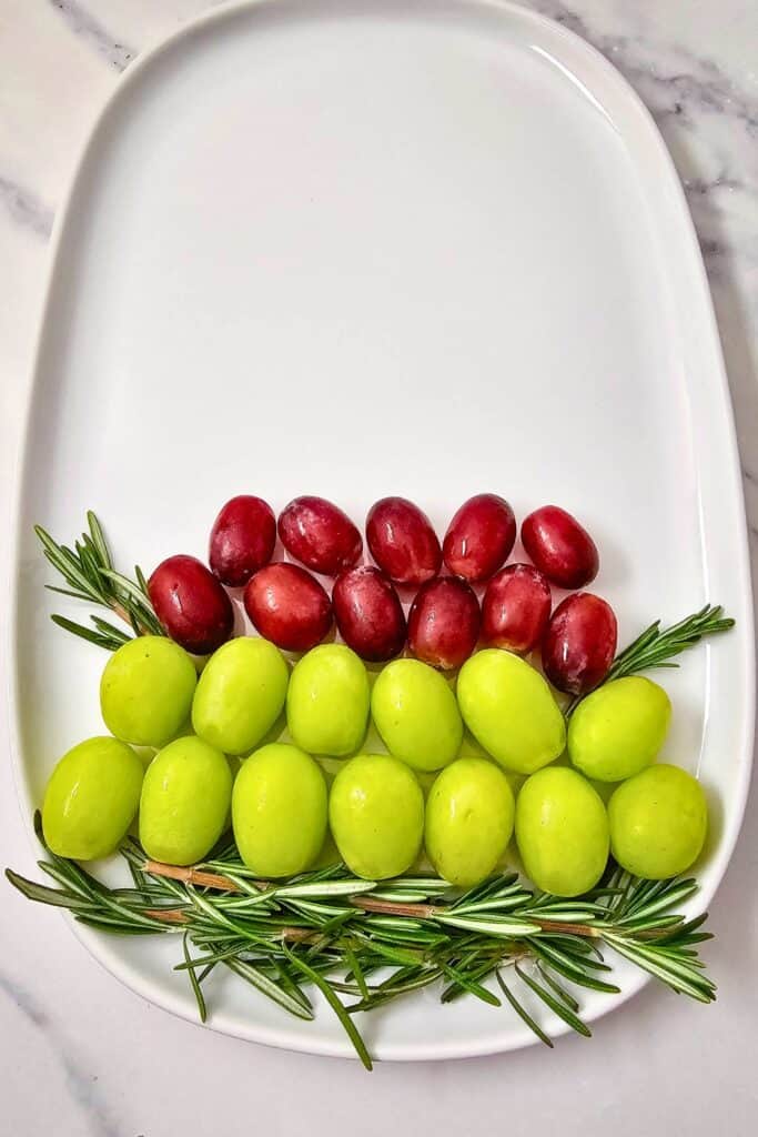 A fruit board idea for Christmas taking shape with neat rows of green and red grapes and fresh rosemary sprigs arranged on a white oval platter to create a festive Christmas tree.