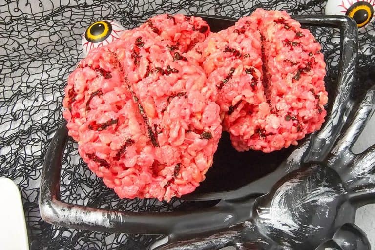 Bright pink rice krispie treats shaped like realistic brains with dark chocolate details for veins, served on a black skeleton hand platter against a spooky netted backdrop with fake eyeballs.