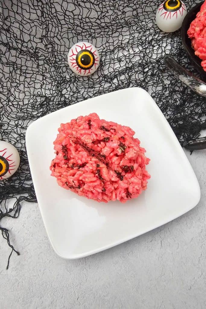 Bright pink rice krispie treat shaped like a brain with dark chocolate vein details, served on a white plate against a spooky black net backdrop with plastic eyeball decorations.