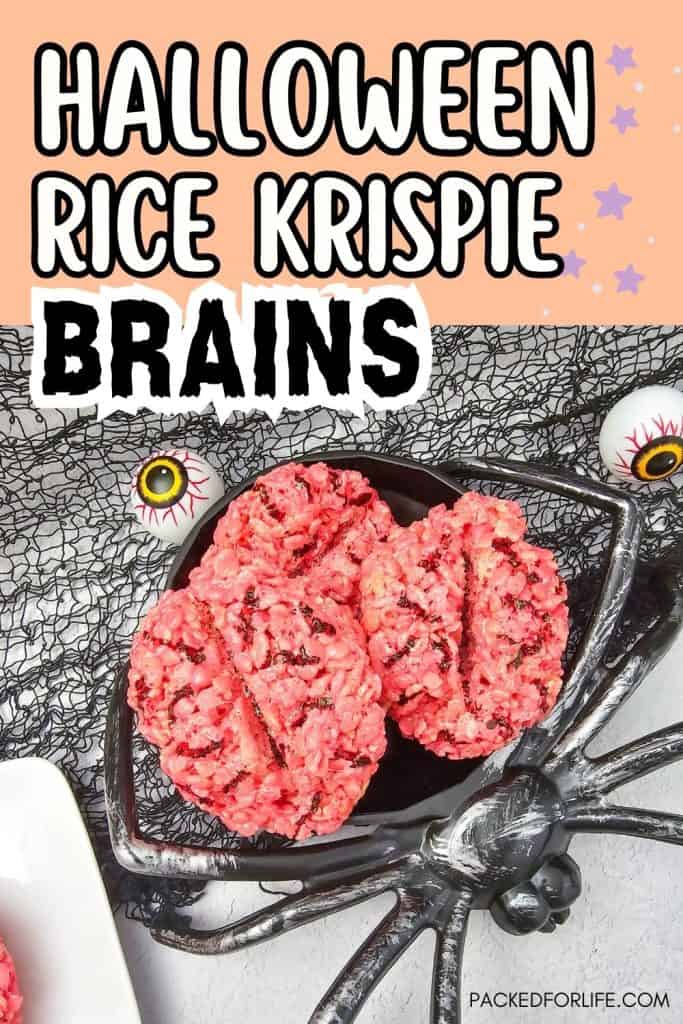 Pink Rice Krispie treats shaped like brains are displayed in a black spider-shaped bowl, with black netting and fake eyeballs for a spooky Halloween backdrop. The top of the image features text that reads “Halloween Rice Krispie Brains” in bold white and black letters.