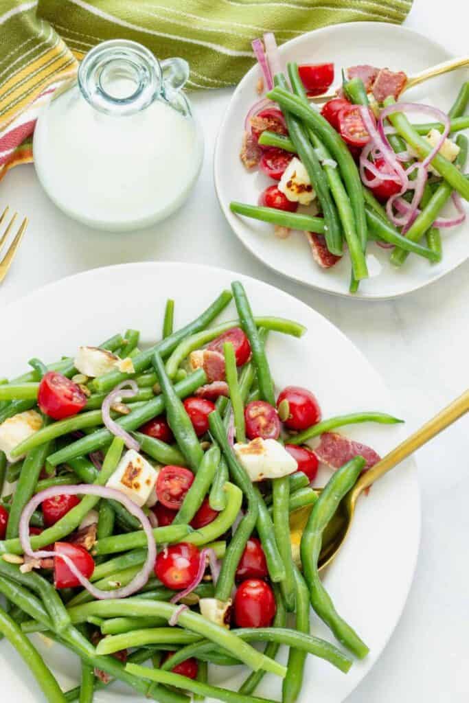 Colorful green bean salad with cherry tomatoes, red onion, grilled cheese cubes, and crispy bacon served on white plates. This fresh and vibrant side salad for summer cookouts is perfect as a light and crunchy side dish.
