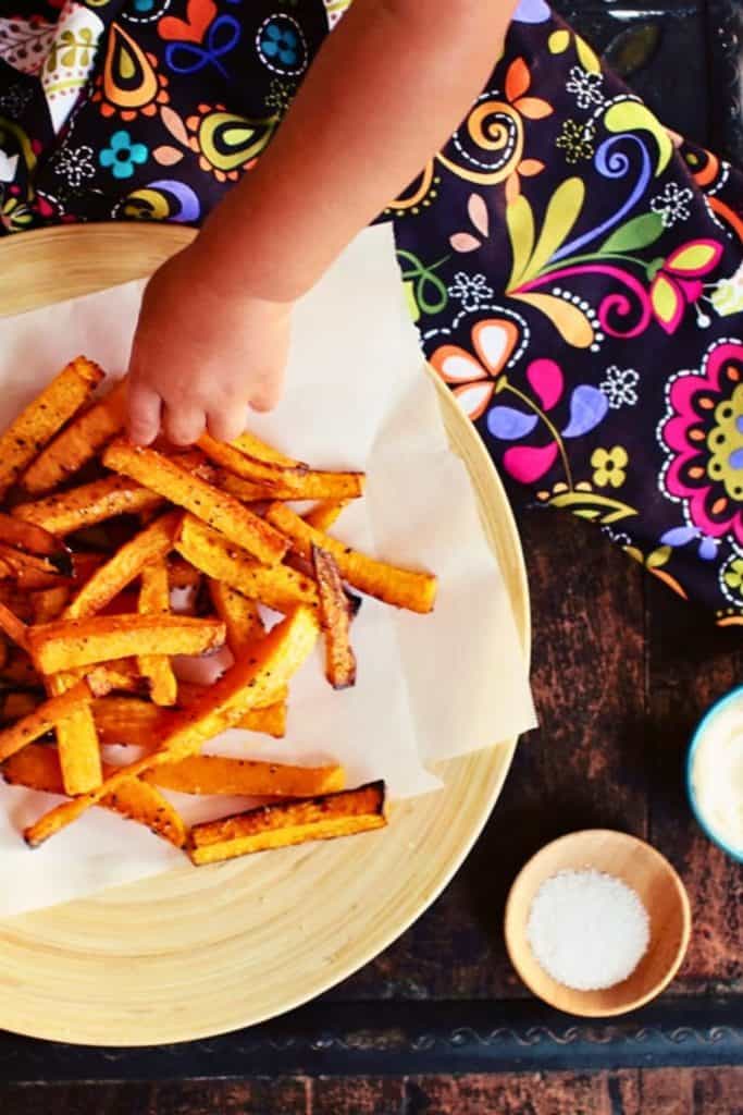A child's hand reaches for a serving of crispy baked butternut squash fries, seasoned and roasted to a golden brown on parchment paper. The fries are served on a wooden plate with a small bowl of sea salt and a colorful patterned cloth in the background.