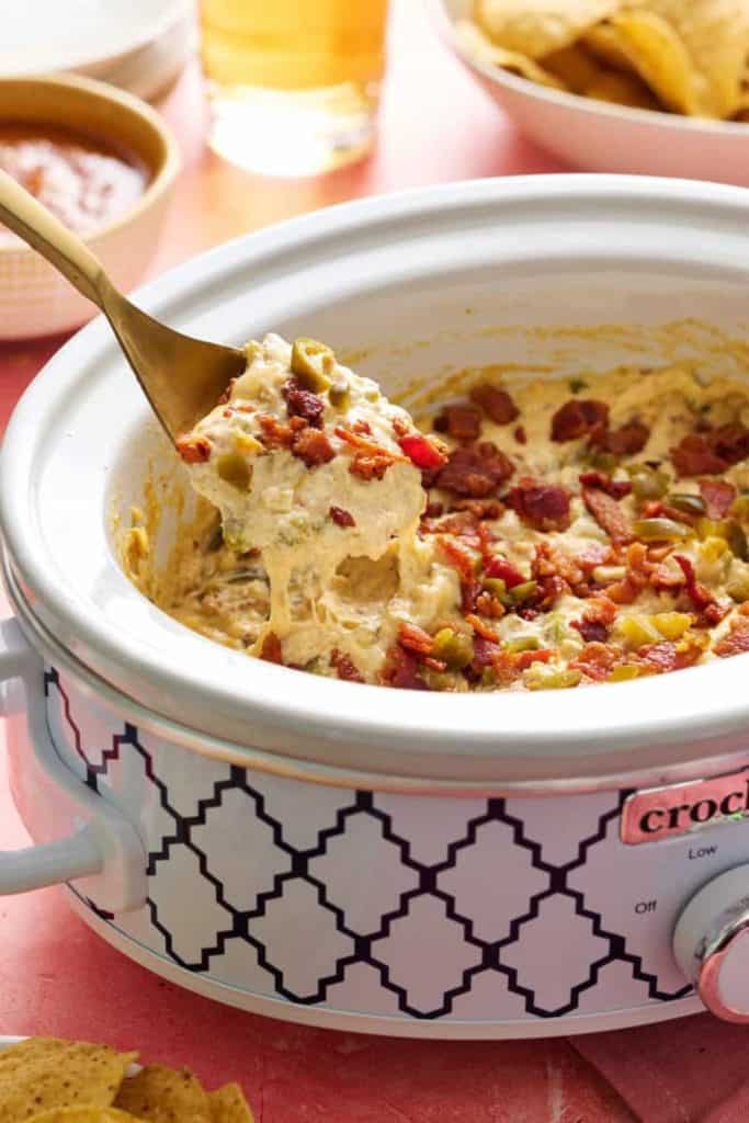 Crock Pot Jalapeño Popper Dip with Bacon in a patterned slow cooker, topped with crispy bacon pieces and diced jalapeños. A spoon lifts a cheesy, creamy scoop.